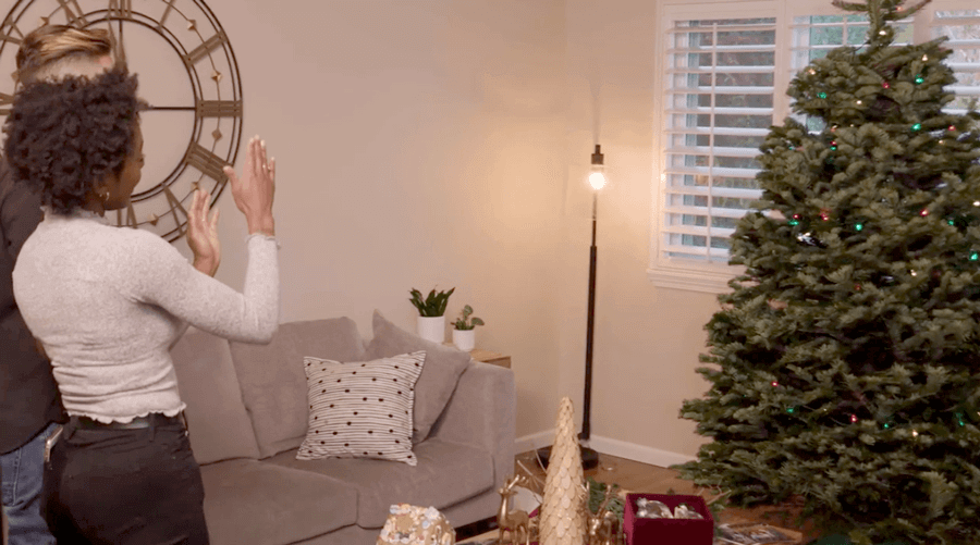A man and a woman standing in a living room trying to see if their Christmas tree is tilted.